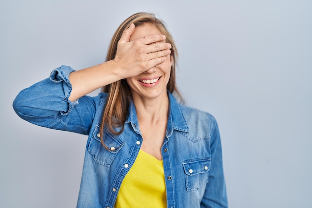 Mujer rubia joven de pie sobre fondo azul sonriendo y riendo con la mano en la cara cubriendo los ojos por concepto ciego sorpresa