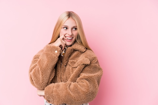 La mujer rubia joven que llevaba un abrigo contra un fondo rosado se relajó pensando en algo que miraba un espacio de la copia.