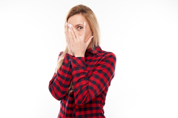 Mujer rubia joven que mira furtivamente a través de los dedos que cubren los ojos con las palmas aisladas en el concepto de la fobia del fondo blanco