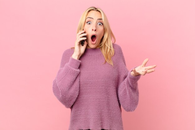 Mujer rubia joven que se siente extremadamente conmocionada y sorprendida y hablando con un teléfono inteligente