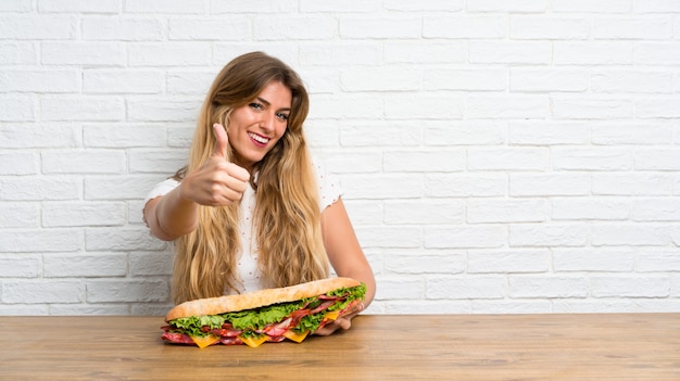 Mujer rubia joven que sostiene un bocadillo grande con el pulgar para arriba