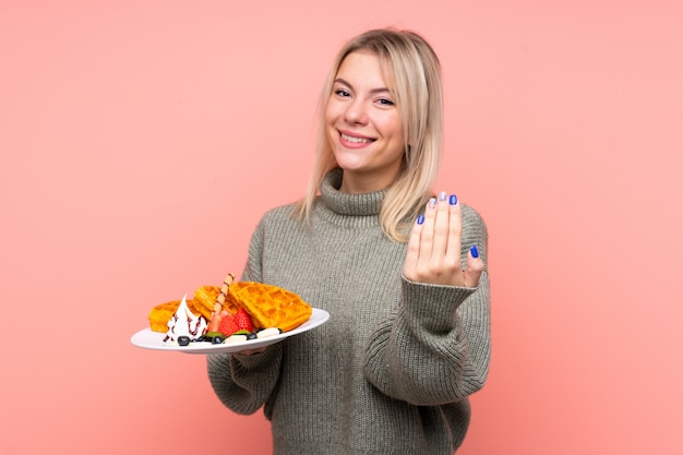 Mujer rubia joven que sostiene las galletas sobre la pared rosada aislada que invita a venir con la mano. Feliz de que hayas venido