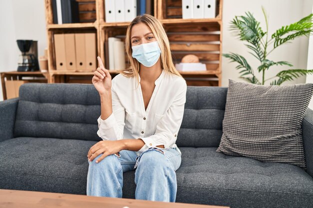 Mujer rubia joven que trata el miedo covid en la terapia sonriendo con una idea o pregunta señalando con el dedo hacia arriba con cara feliz, número uno