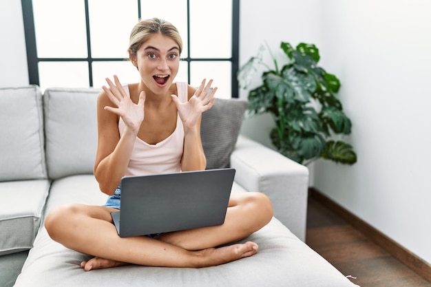 Mujer rubia joven que usa una computadora portátil en casa sentada en el sofá celebrando loca y sorprendida por el éxito con los brazos levantados y los ojos abiertos gritando el concepto de ganador emocionado