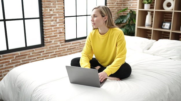 Mujer rubia joven que usa la computadora portátil que se sienta en cama en el dormitorio