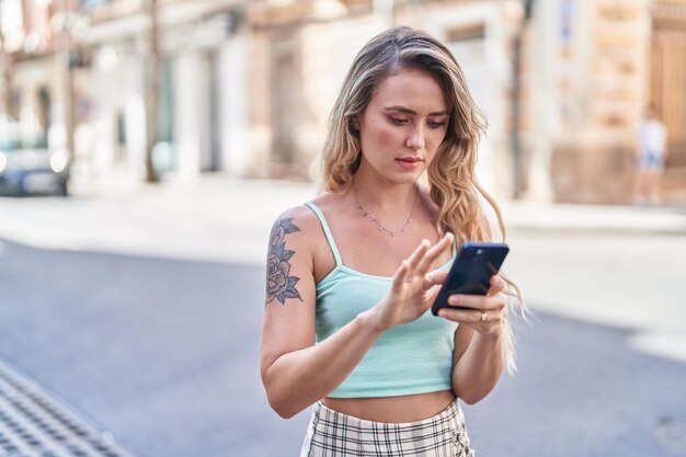 Mujer rubia joven que usa el teléfono inteligente con expresión seria en la calle