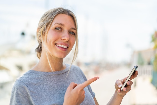 Mujer rubia joven que usa el teléfono móvil al aire libre y lo señala