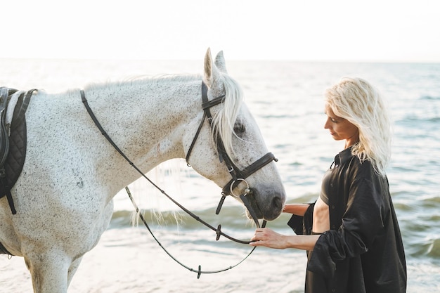 Mujer rubia joven en ropa negra montando caballo blanco sobre fondo marino