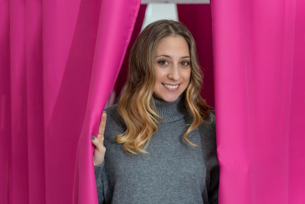 Mujer rubia joven saliendo de detrás de cortinas rosas en un vestidor Ella está sonriendo