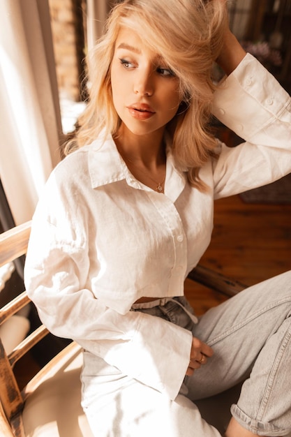 Mujer rubia joven sexy en una camisa de lino blanco y jeans se sienta y descansa en una silla de madera en un día soleado en la habitación