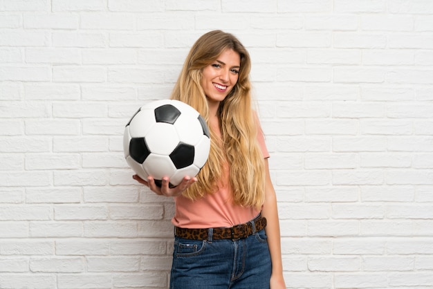 Mujer rubia joven sobre la pared de ladrillo blanca que sostiene un balón de fútbol