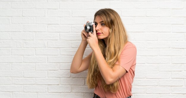 Mujer rubia joven sobre la pared de ladrillo blanca que sostiene una cámara