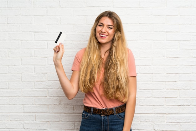 Mujer rubia joven sobre la pared de ladrillo blanca que sostiene una tarjeta de crédito