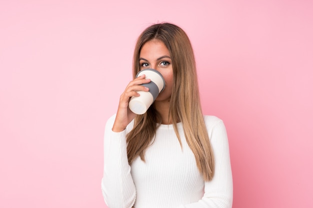 Mujer rubia joven sobre la pared rosada aislada que sostiene y que bebe un café para llevar