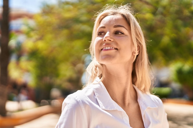 Mujer rubia joven sonriendo confiada de pie en el parque