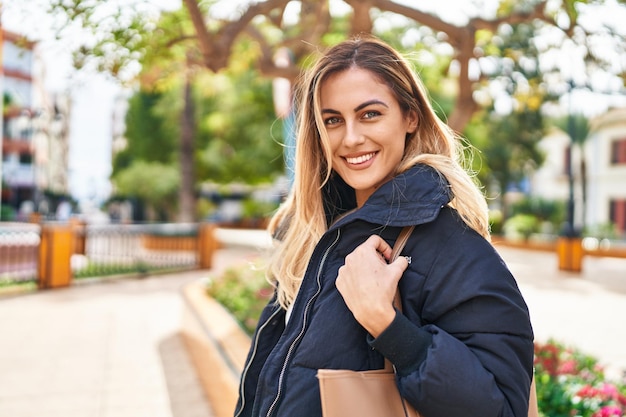 Mujer rubia joven sonriendo confiada de pie en el parque