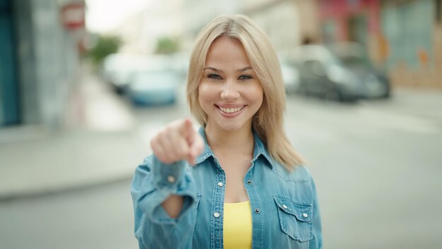 Mujer rubia joven sonriendo confiada señalando con el dedo en la calle