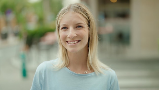 Mujer rubia joven sonriendo segura de pie en la calle