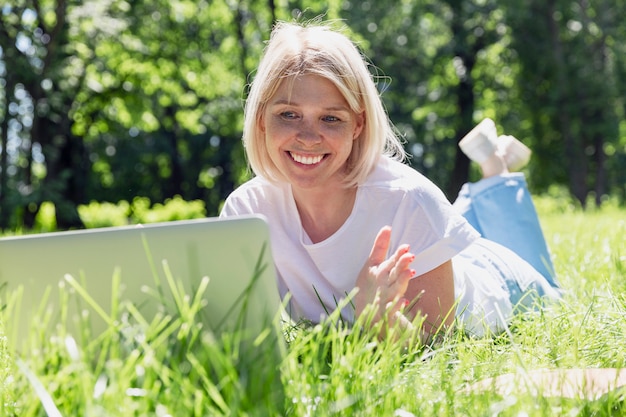 Una mujer rubia joven sonriente miente con una computadora portátil en la hierba en un parque en un día soleado de verano. Blogging, comunicación en línea y redes sociales.