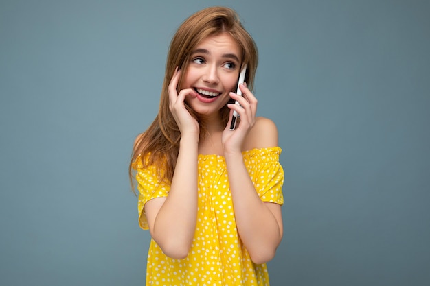 Mujer rubia joven sonriente positiva atractiva atractiva pensativa que lleva el vestido amarillo elegante del verano que se encuentra aislado sobre la pared azul que sostiene y que habla por teléfono móvil que mira hacia el lado.