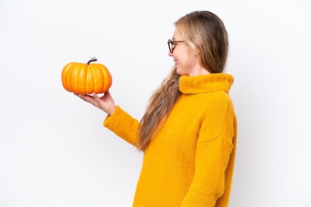 Mujer rubia joven sosteniendo una calabaza aislada sobre fondo blanco con expresión feliz