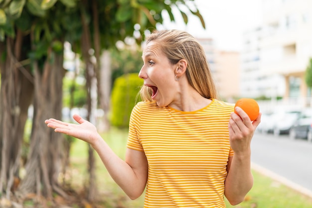Mujer rubia joven sosteniendo una naranja al aire libre con expresión facial de sorpresa