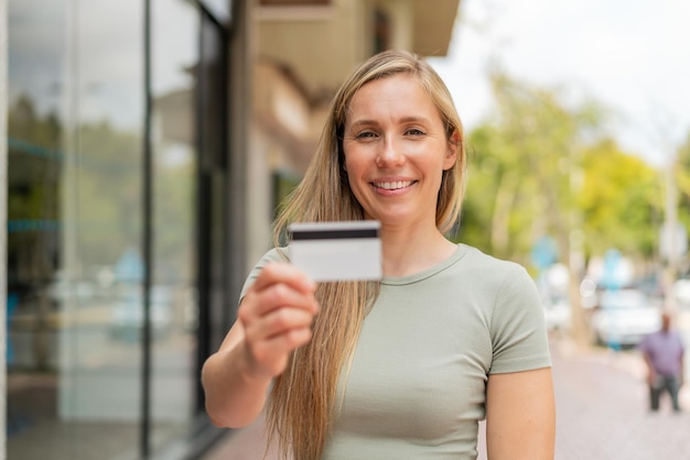 Mujer rubia joven sosteniendo una tarjeta de crédito al aire libre con expresión feliz