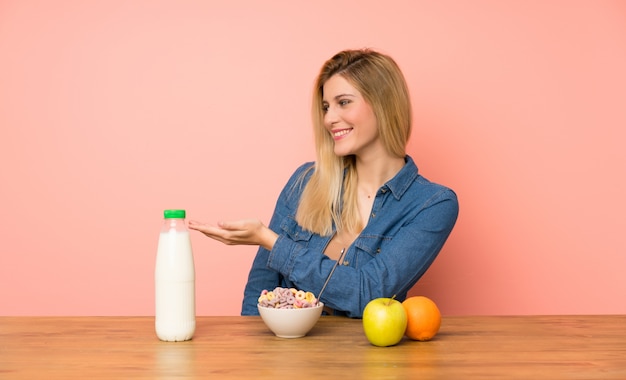 Mujer rubia joven con un tazón de cereales que extiende las manos hacia un lado para invitar a venir