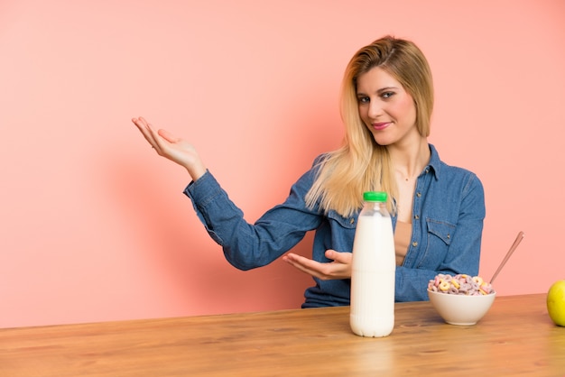 Foto mujer rubia joven con un tazón de cereales que extiende las manos hacia un lado para invitar a venir