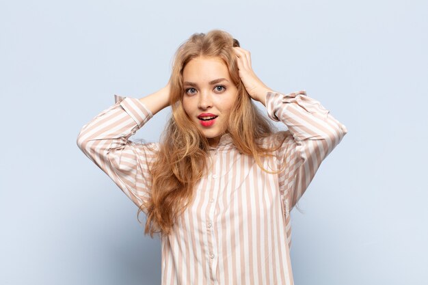 Mujer rubia levantando las manos a la cabeza, con la boca abierta, sintiéndose extremadamente afortunada, sorprendida, emocionada y feliz