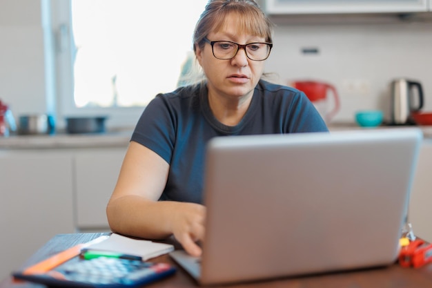 Mujer rubia madura cansada independiente que trabaja desde casa usando una computadora portátil sentada en la cocina