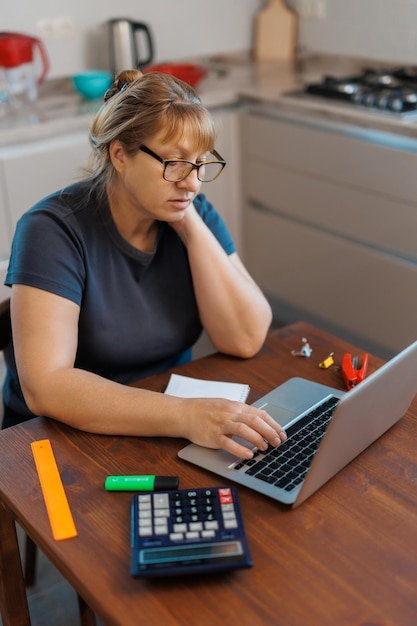 Mujer rubia madura cansada independiente que trabaja desde casa usando una computadora portátil sentada en la cocina