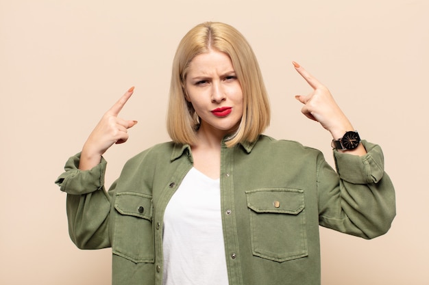 Mujer rubia con mala actitud que parece orgullosa y agresiva, apuntando hacia arriba o haciendo letreros divertidos con las manos