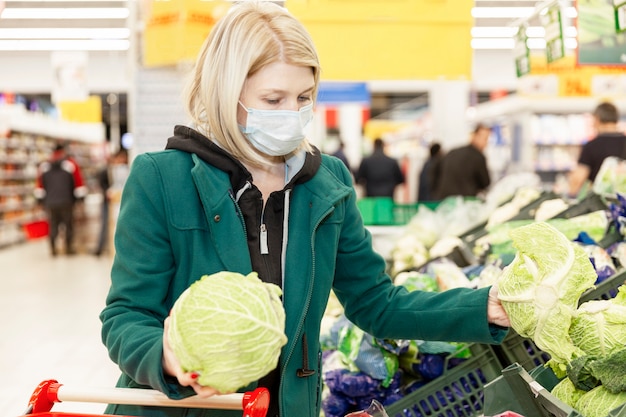 La mujer rubia en una máscara médica elige verduras en un supermercado. Autoaislamiento en una pandemia.