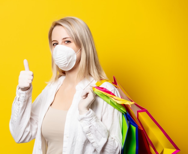 Mujer rubia en mascarilla sosteniendo bolsas de compras en la pared amarilla