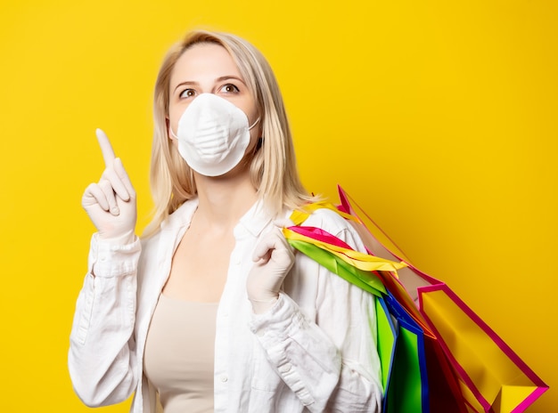 Foto mujer rubia en mascarilla sosteniendo bolsas de compras en la pared amarilla