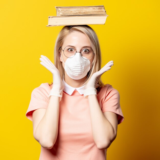 Mujer rubia en mascarilla y vestido rosa sosteniendo un libro en la pared amarilla