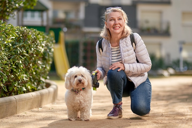 Mujer rubia de mediana edad caminando con un perro blanco esponjoso en la ciudad de verano.