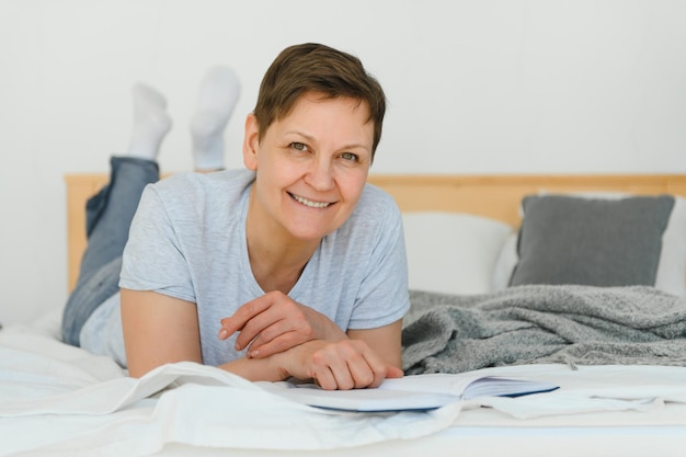 Mujer rubia de mediana edad leyendo un libro acostado en la cama en casa