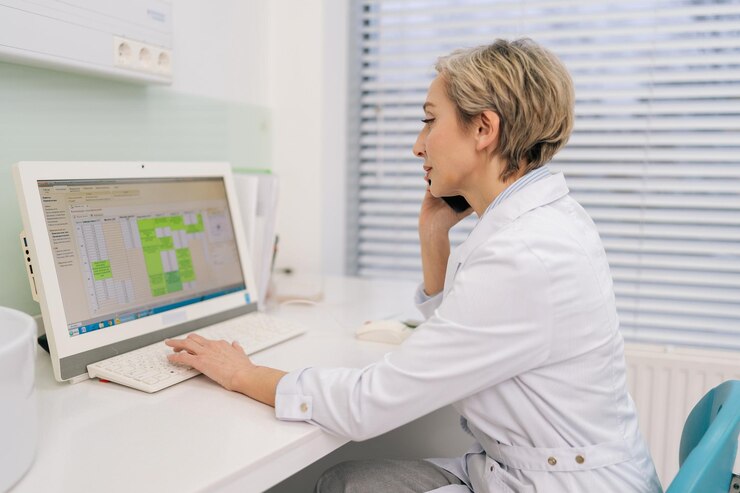 Foto mujer rubia de mediana edad, médico general, sentada en el escritorio de la oficina de la clínica mirando la pantalla del portátil