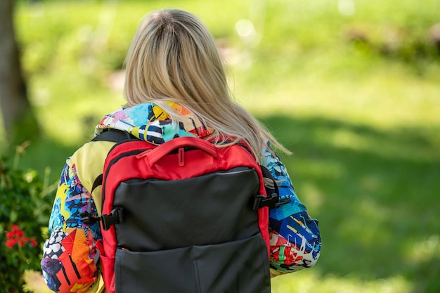 Mujer rubia de mediana edad con ropa brillante con una mochila en el parque en un día soleado vista trasera