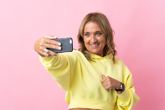 Mujer rubia de mediana edad sobre fondo rosa aislado haciendo un selfie con teléfono móvil