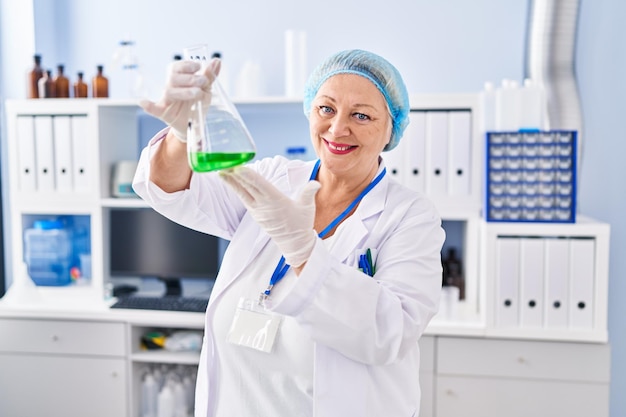 Mujer rubia de mediana edad con uniforme científico sosteniendo tubo de ensayo en el laboratorio