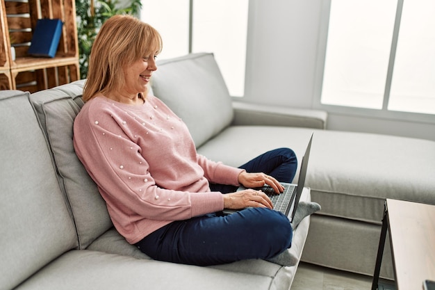 Mujer rubia de mediana edad usando una laptop sentada en el sofá en casa