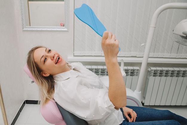 Mujer rubia mira en el espejo a sus dientes, sentada en una silla. Niña sonriente en el consultorio del dentista