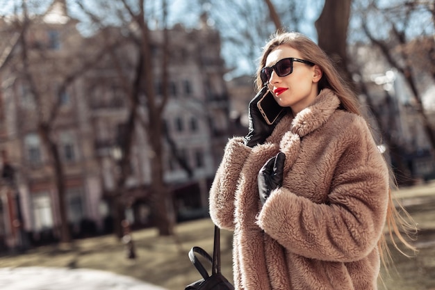 Mujer rubia de moda atractiva joven vestida con abrigo de piel y gafas de sol hablando por teléfono en la ciudad