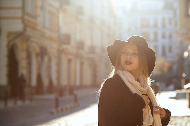 Mujer rubia de moda con sombrero, bufanda y abrigo, caminando por la calle vieja bajo los rayos del sol. Espacio para texto