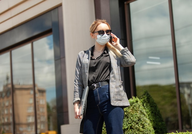 Foto mujer rubia de negocios con una máscara médica y gafas de sol hablando por teléfono y sostiene una computadora portátil en sus manos en la ciudad