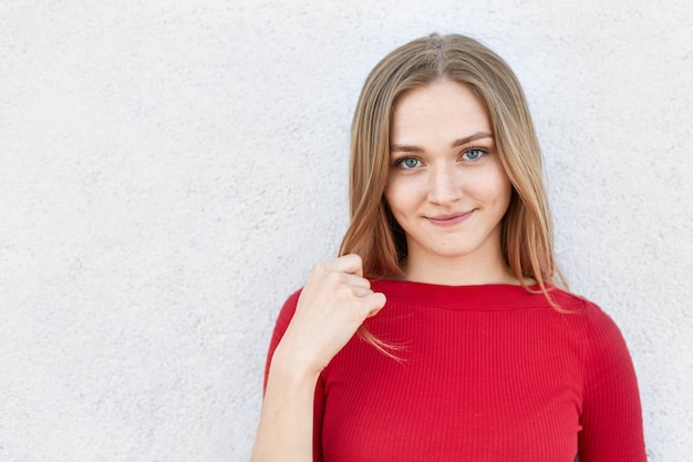 Mujer rubia con ojos azules y cara pecosa mirando seriamente a la cámara mientras está de pie cerca de la pared blanca