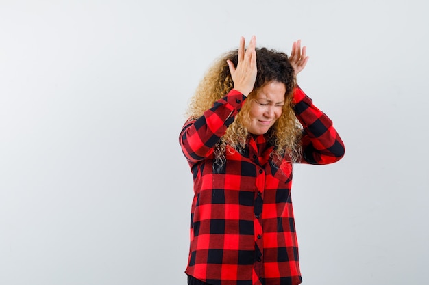 Foto mujer rubia con pelo rizado en camisa a cuadros manteniendo las manos en la cabeza y mirando deprimido, vista frontal.